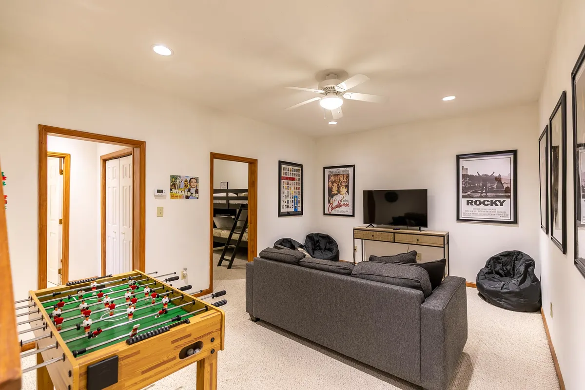 The image features a vibrant game room with a foosball table as the centerpiece, a comfortable sofa for lounging, a large TV, and playful posters on the wall. The atmosphere is casual and inviting, ideal for entertaining groups or families.