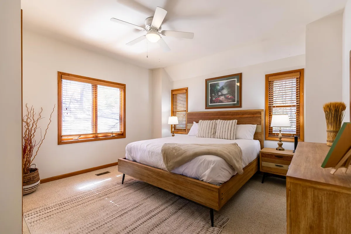 This tranquil bedroom features a cozy bed, nightstands, windows, and tasteful artwork, illuminated by natural light. The setting emphasizes comfort and a restful atmosphere, ideal for unwinding after a day of seasonal mountain adventures.