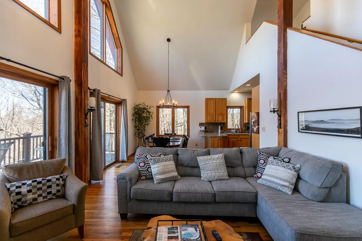 This image showcases a spacious sectional sofa set beneath large windows with sunset views, exposed wooden beams, and a visible kitchen area. The overall atmosphere is cozy and inviting, suggesting a place where guests and their pets can unwind after a day of outdoor activities.