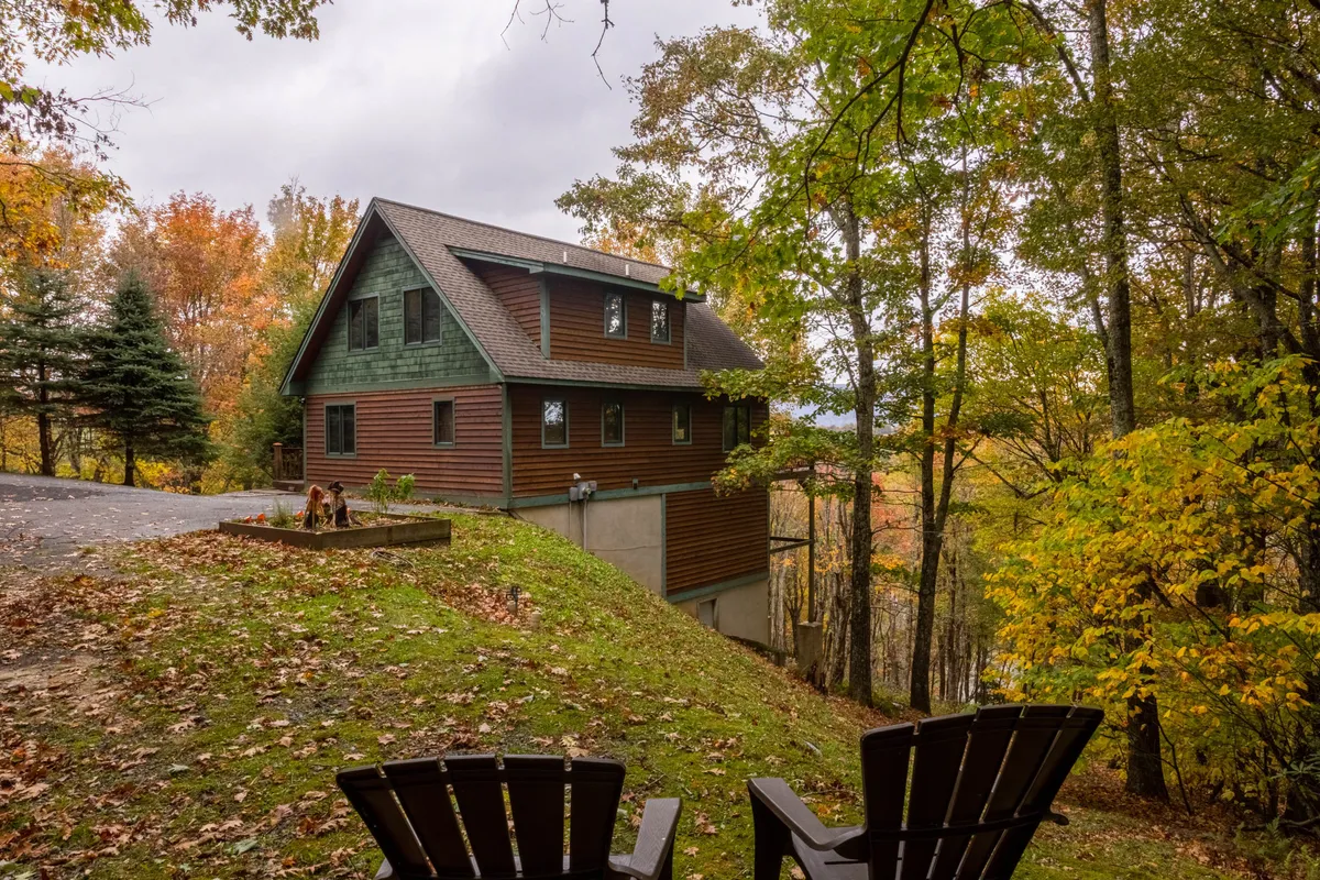 A serene view of the home's exterior, with the house nestled among mature trees. Comfortable chairs invite guests to relax on the porch, and the lush greenery underscores the peaceful setting. The foreground features inviting outdoor seating perfect for gathering or enjoying quiet moments.