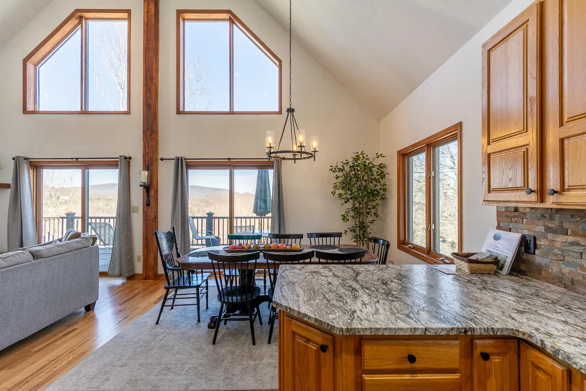A spacious open-concept area featuring a dining table set for a group, large windows that frame beautiful outdoor views, a cozy sofa, and a kitchen island beneath a striking chandelier. The scene is lively, inviting, and ideal for socializing or hosting group meals.