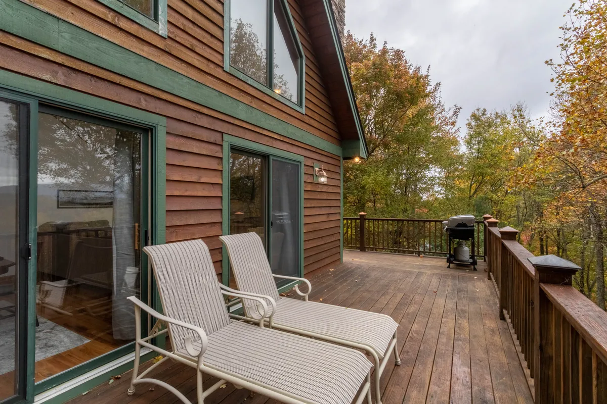 An exterior shot of the deck, with comfortable seating, a grill, and surrounding trees visible. The scene suggests a seamless transition from the interior living space to outdoor al fresco dining and relaxation.