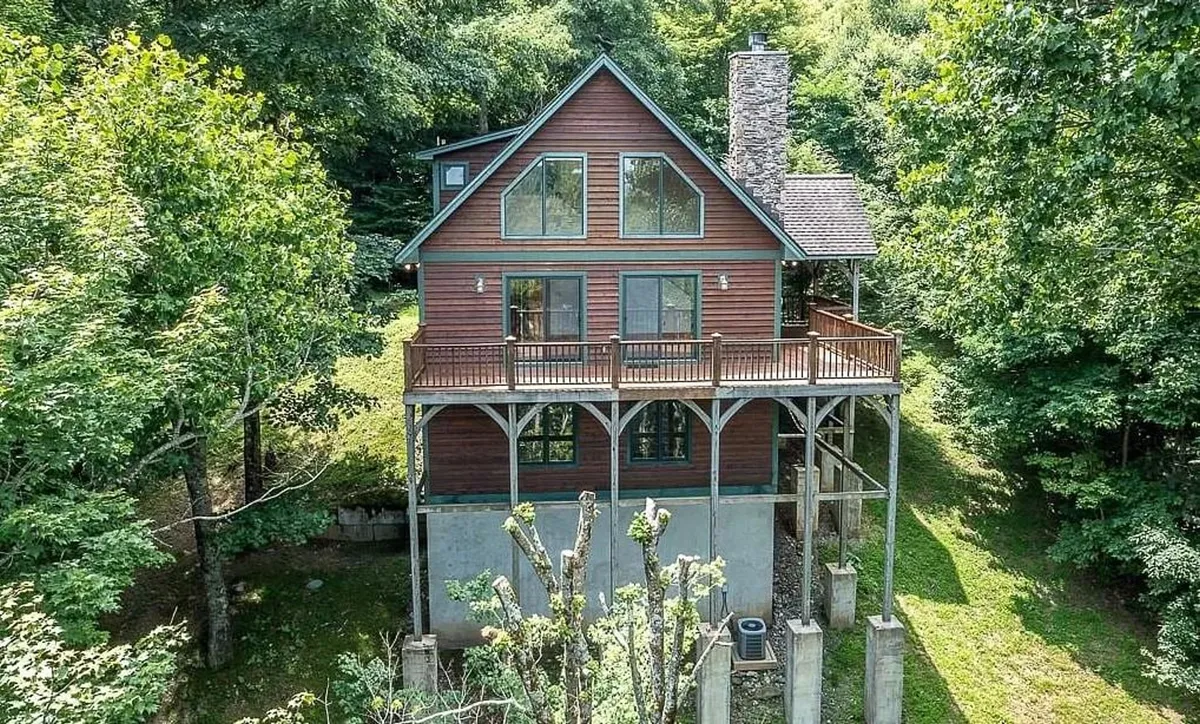 The image captures the exterior of a welcoming mountain home nestled among tall trees, featuring a spacious wooden deck perfect for relaxing or family gatherings, a striking stone chimney, and large windows that invite natural light and offer breathtaking views of the surrounding landscape.