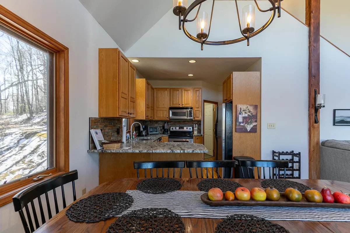 An inviting dining area centered around a table adorned with a fruit centerpiece, accented by a modern chandelier above and a roomy kitchen island nearby. The setting feels perfect for communal cooking, sharing meals, and celebrating together.
