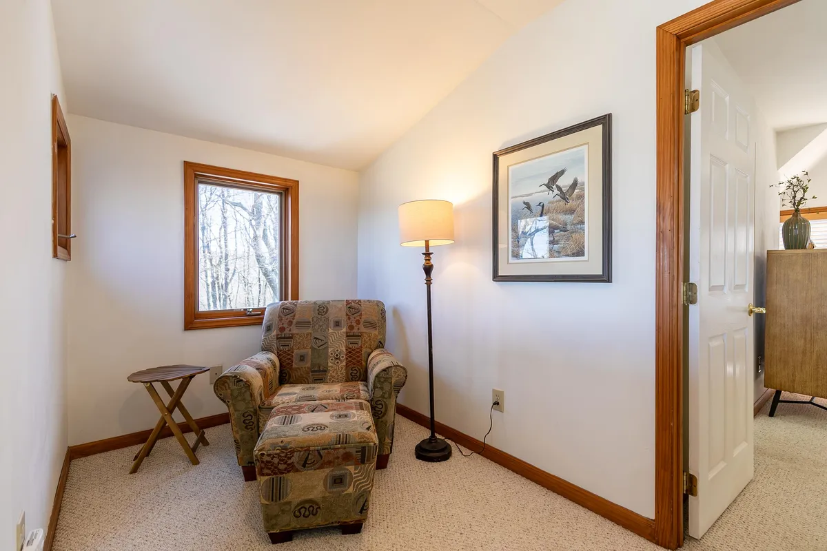 A serene reading nook featuring a plush armchair beside a window, a lamp for soft illumination, and a side table—perfect for a quiet escape or enjoying nature views. Framed artwork and gentle light enhance the peaceful mood.