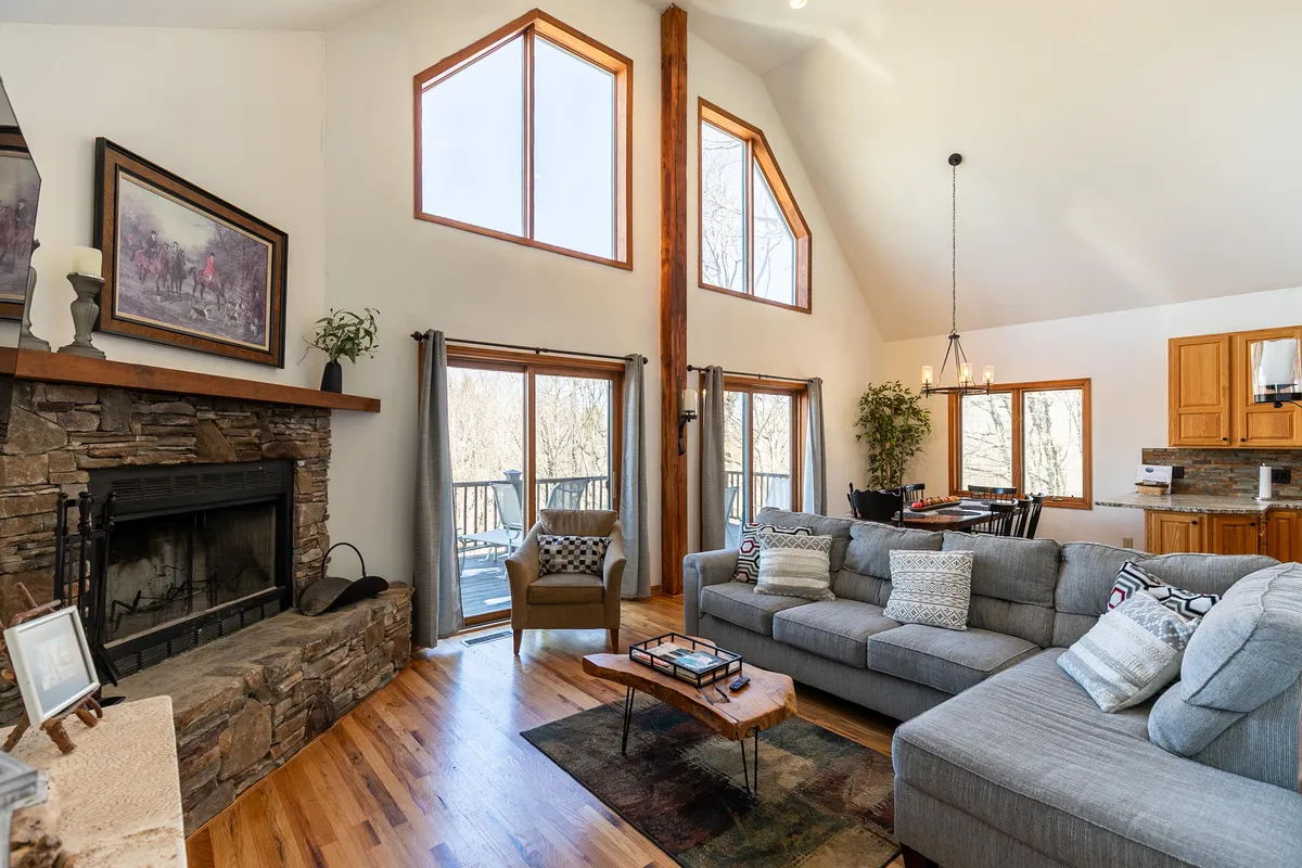 This image showcases the great room with a cozy sofa, a wood-burning fireplace as the focal point, expansive windows that frame forested views, and a connected dining area. The scene exudes warmth and invites both relaxation and socializing, with plenty of natural light highlighting the open-concept design.